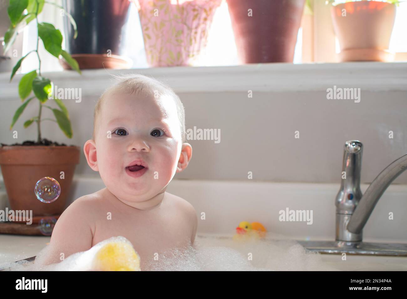 Baby taking bath in the kitchen sink. Child playing with foam and soap