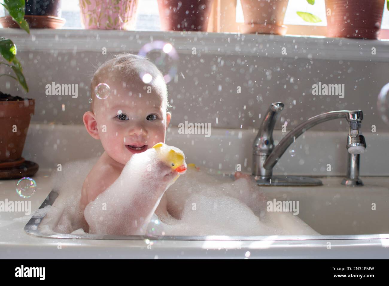 Baby taking bath in the kitchen sink. Child playing with foam and soap