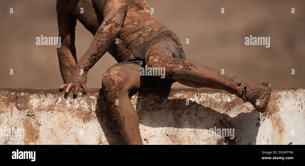 Mud race runners running, participant jump over a concrete wall obstacle Stock Photo