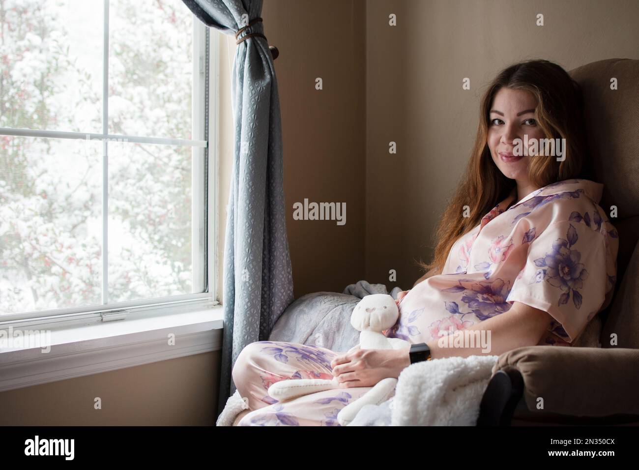 A pregnant caucasian young woman sitting in the rocking chair near the window smiling and looking at her belly Stock Photo