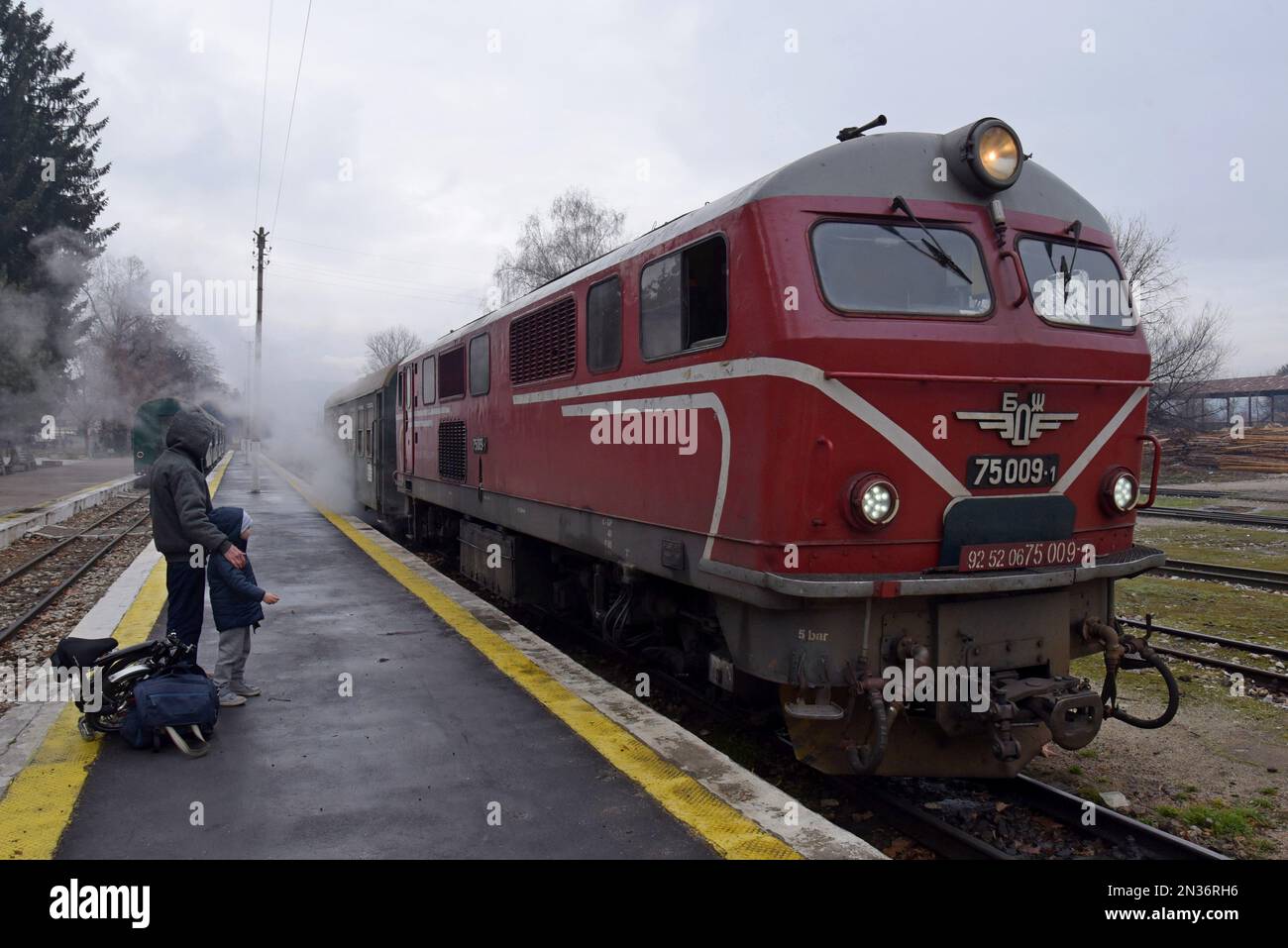 Passengers watch a 1960's built Henschel diesel loco arriving at Velingrad station, Rhodope narrow gauge railway, Bulgaria steam leaking from heating - Stock Image