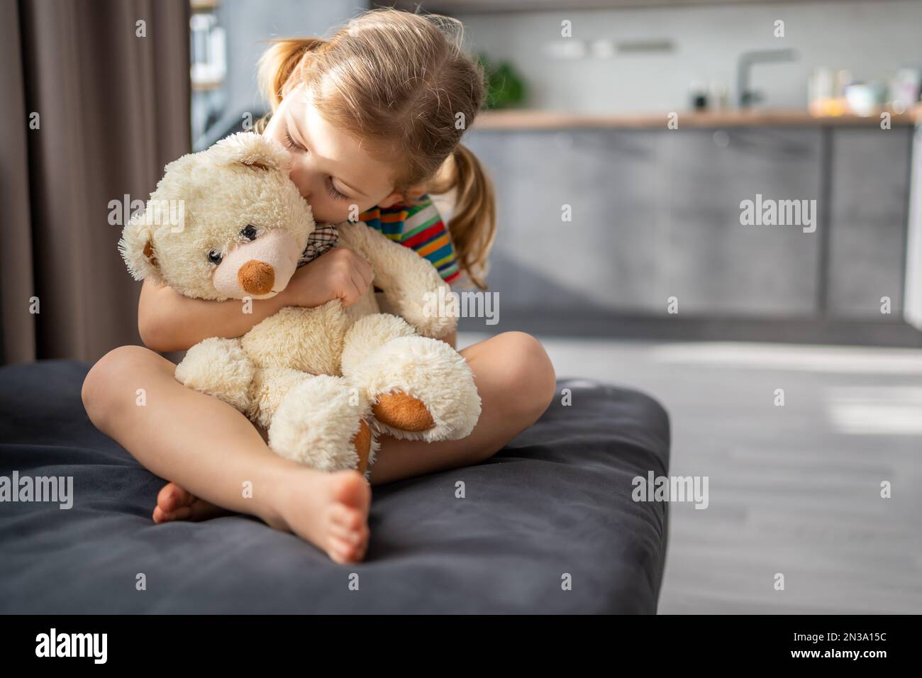 Cute little girl hugging teddy bear while sitting on sofa at home Stock ...