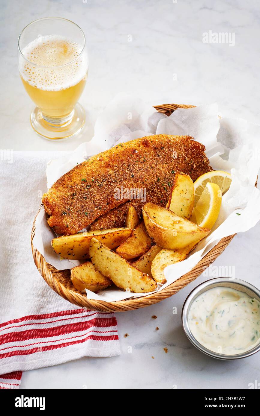 Breaded tilapia with potato wedges in a basket and tarter sauce and a glass of beer Stock Photo