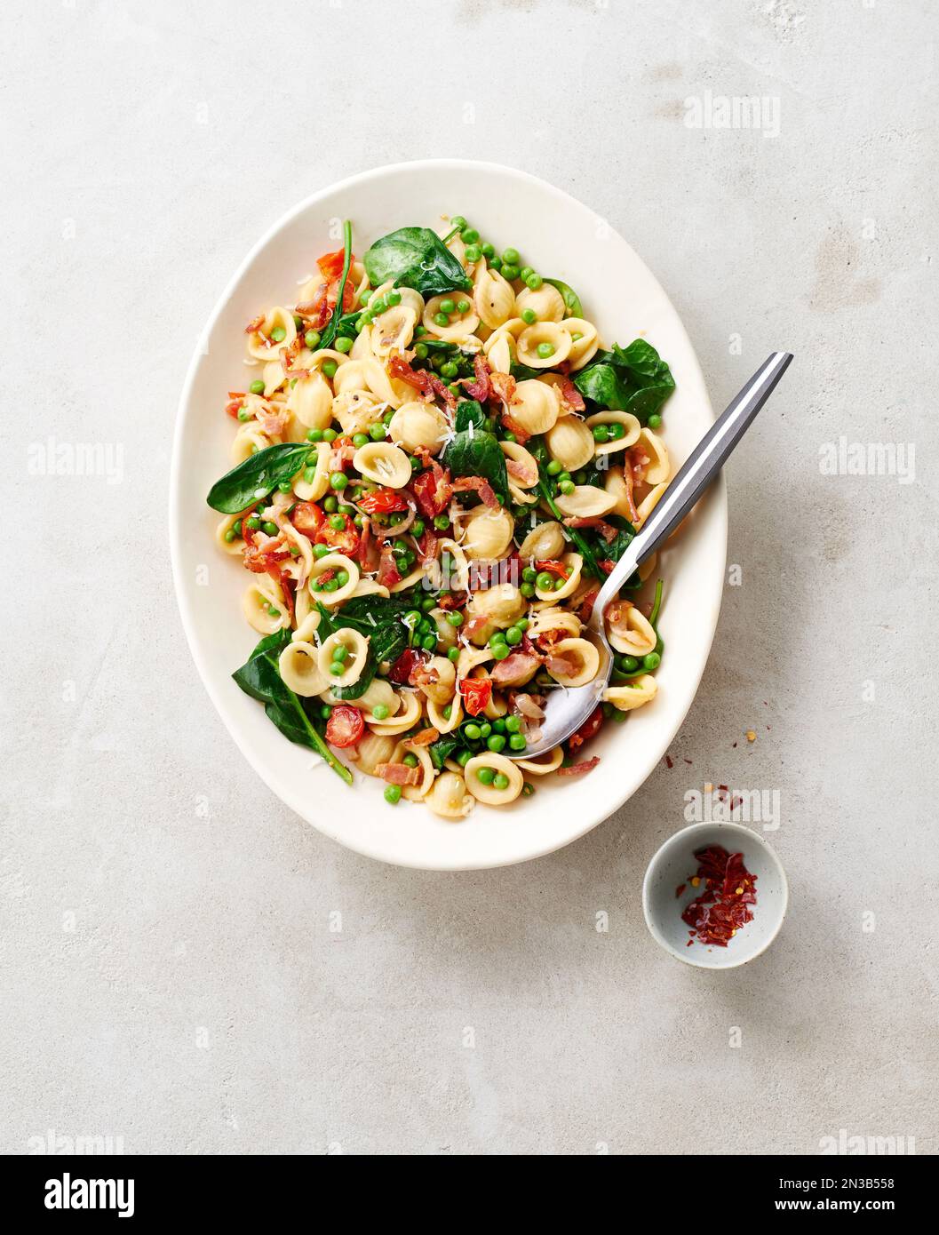 Platter of orecchiette pasta with peas, bacon, spinach and tomatoes and chili flakes in a small bowl on a beige background Stock Photo