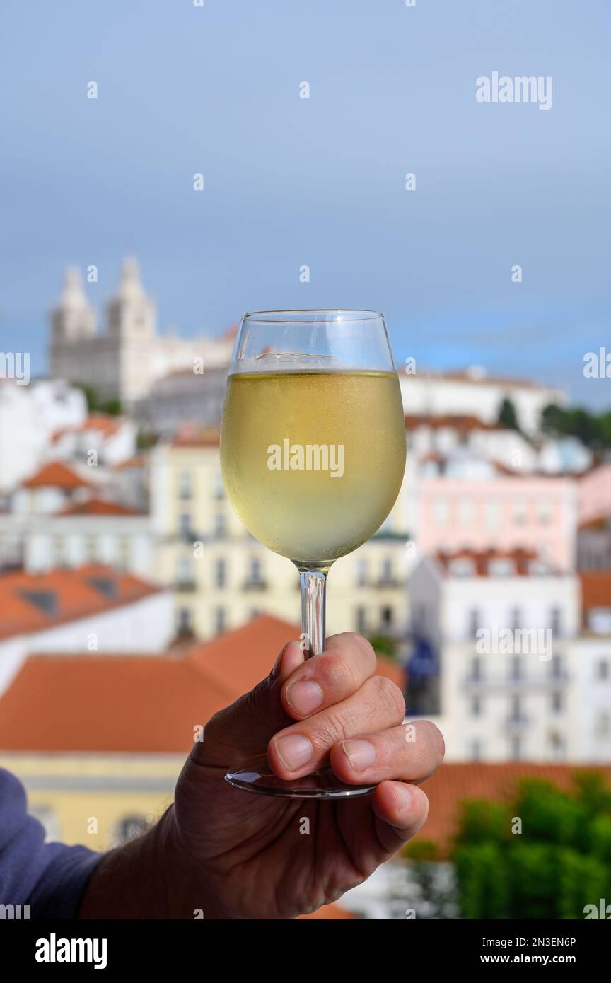 Hand with glass of cold white Portuguese wine in outdoor cafe at view point on colorful old part of Lisbon city, Portugal Stock Photo