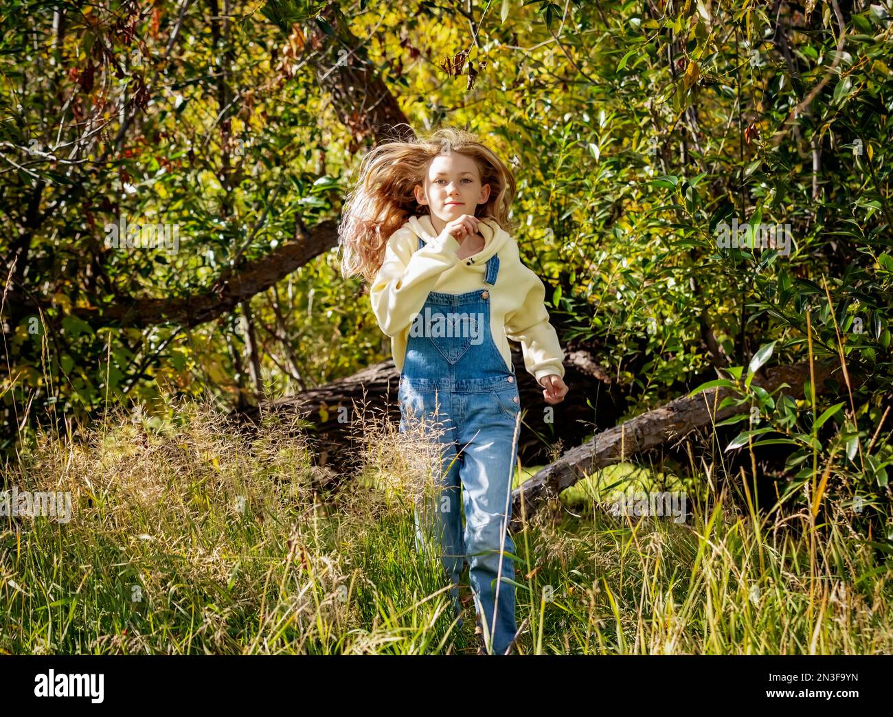 Young girl with long blond hair running out of the forest towards the camera during a warm fall day in a city park; St. Albert, Alberta, Canada Stock Photo