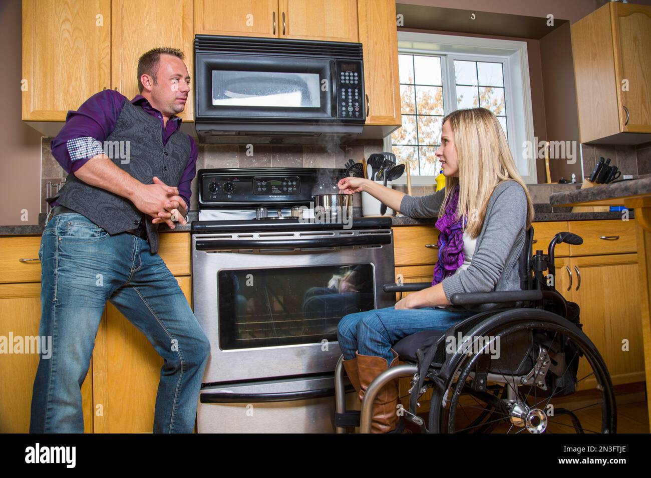 Husband and wife, who has a disability and is in a wheelchair, having a discussion in their kitchen while she is cooking Stock Photo