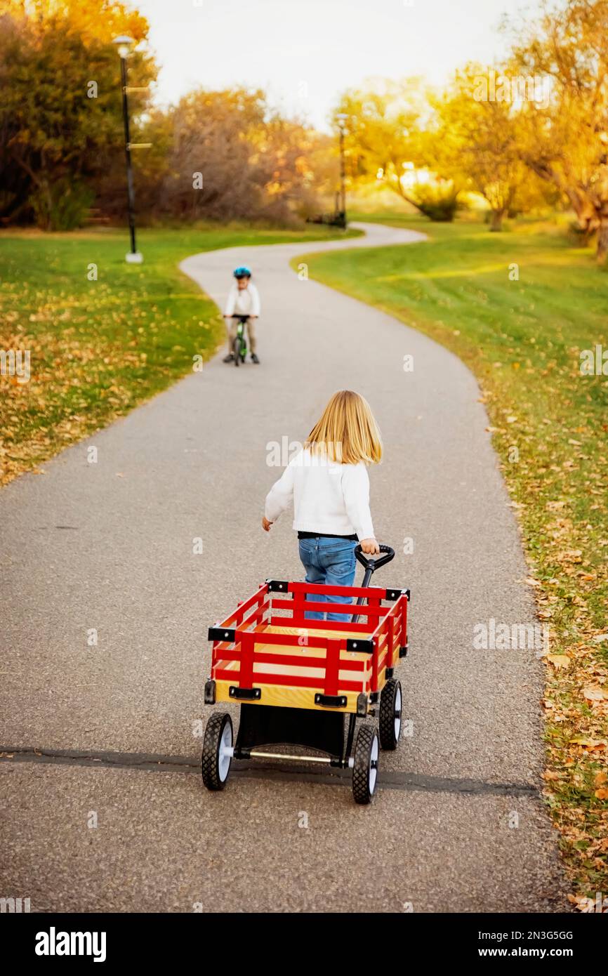 Young girl pulling her wagon along a path in a city park during the fall season with her brother riding a bicycle towards her Stock Photo