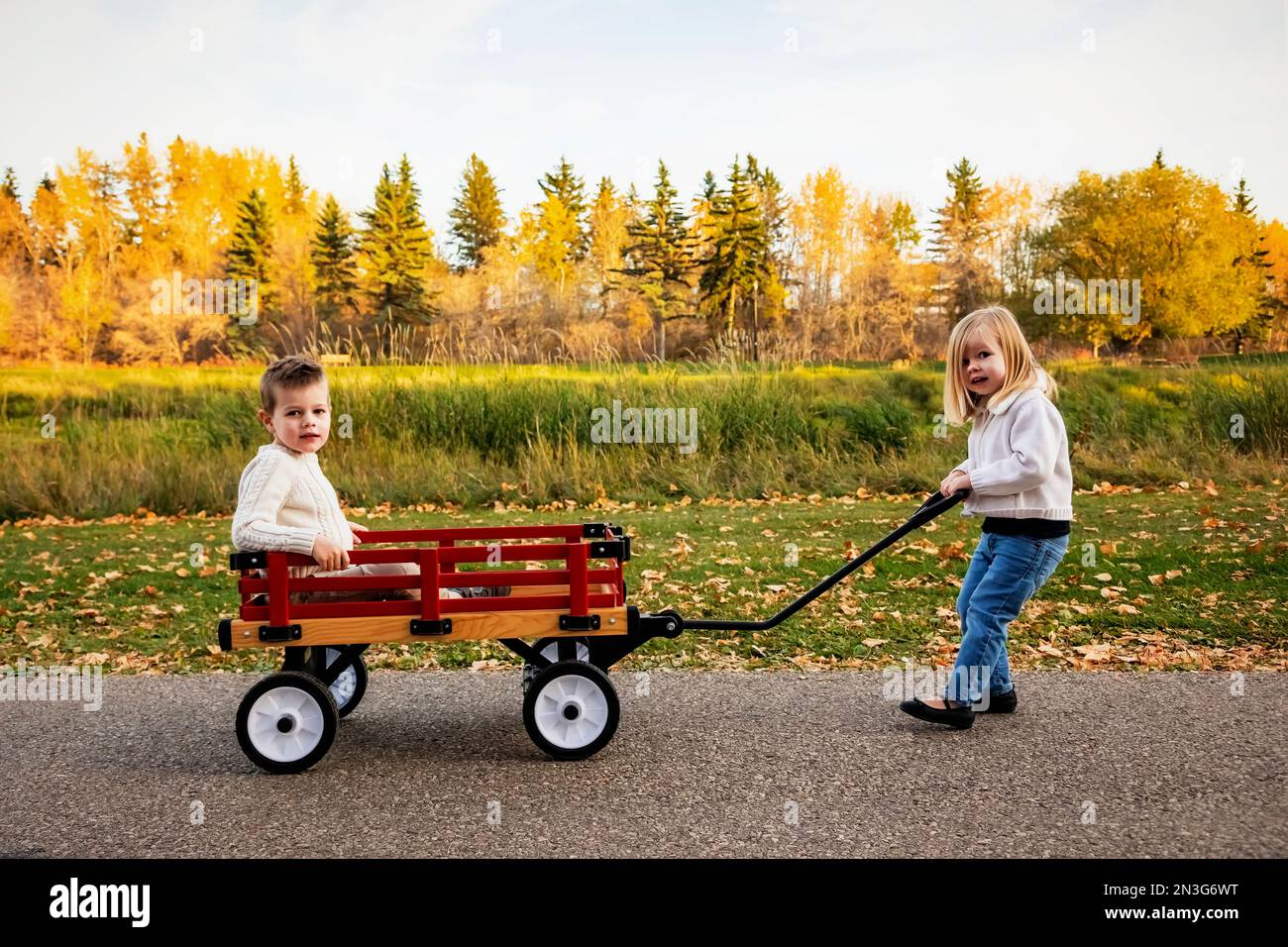 Young girl pulling her brother in a wagon at a city park along a river ...