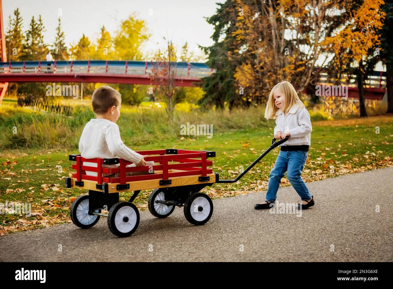 Young girl pulling her brother in a wagon at a city park along a river ...