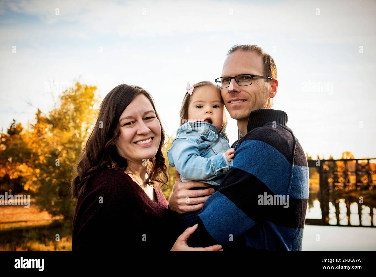 Family portrait while standing on a bridge over a river in a city park during the fall seaon and their baby daughter has Down Syndrome Stock Photo