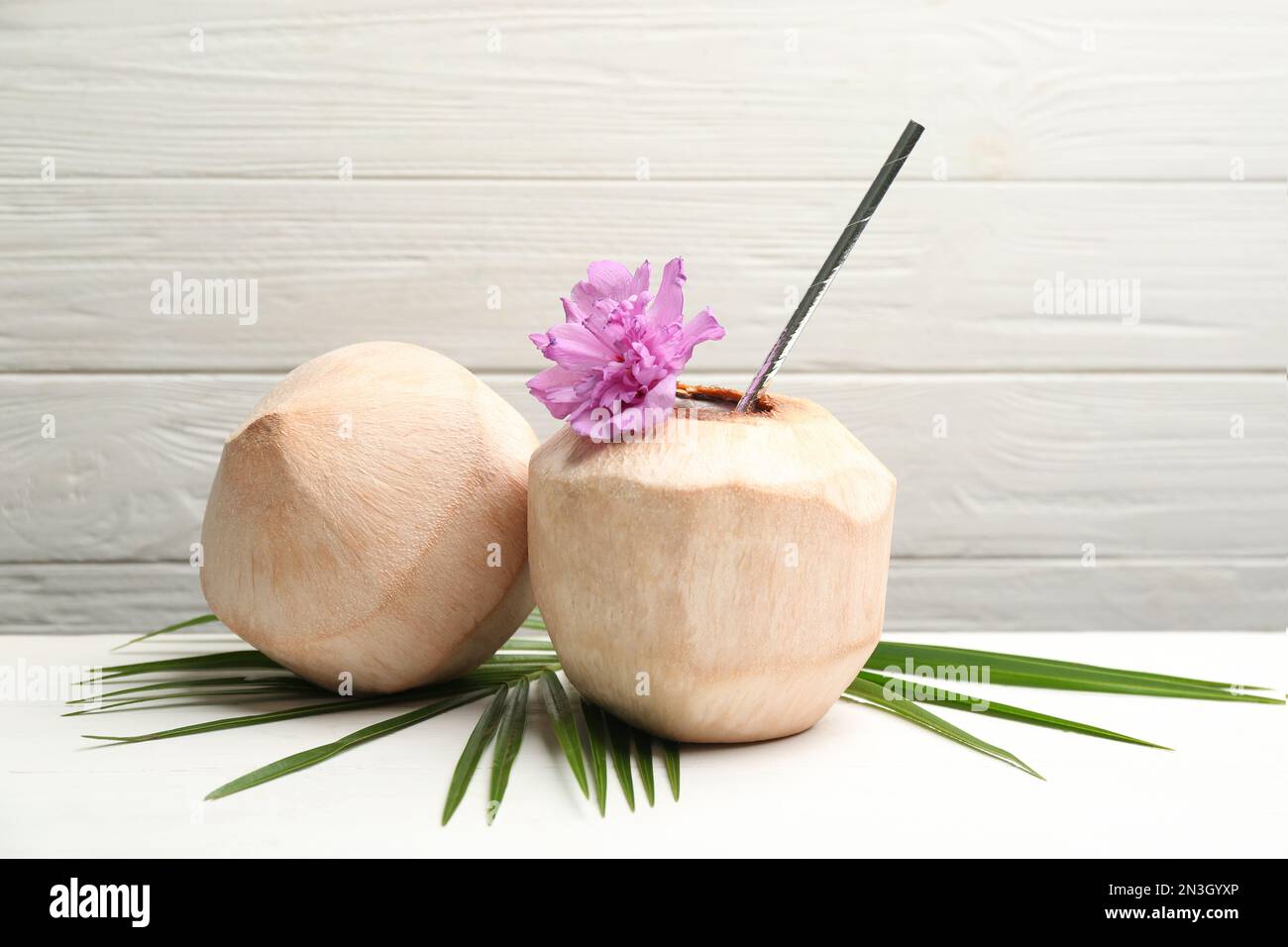Young peeled coconuts with straw, palm leaf and flower on white wooden table Stock Photo