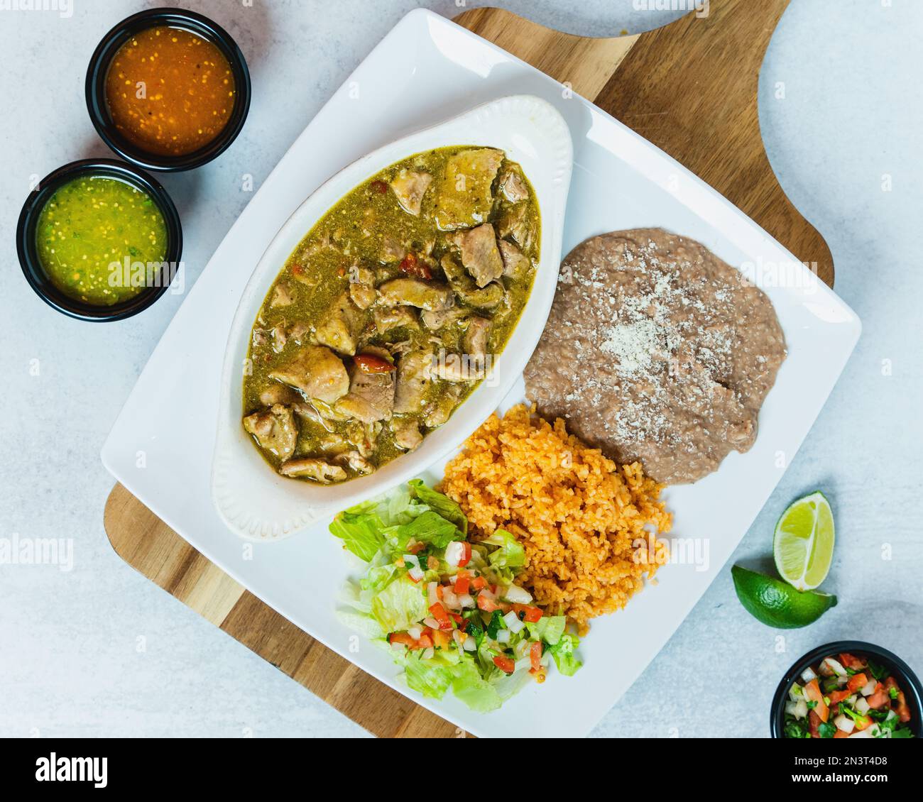 A top view of chile verde plate on a wooden board with refried beans, rice, salsa, and lime Stock Photo
