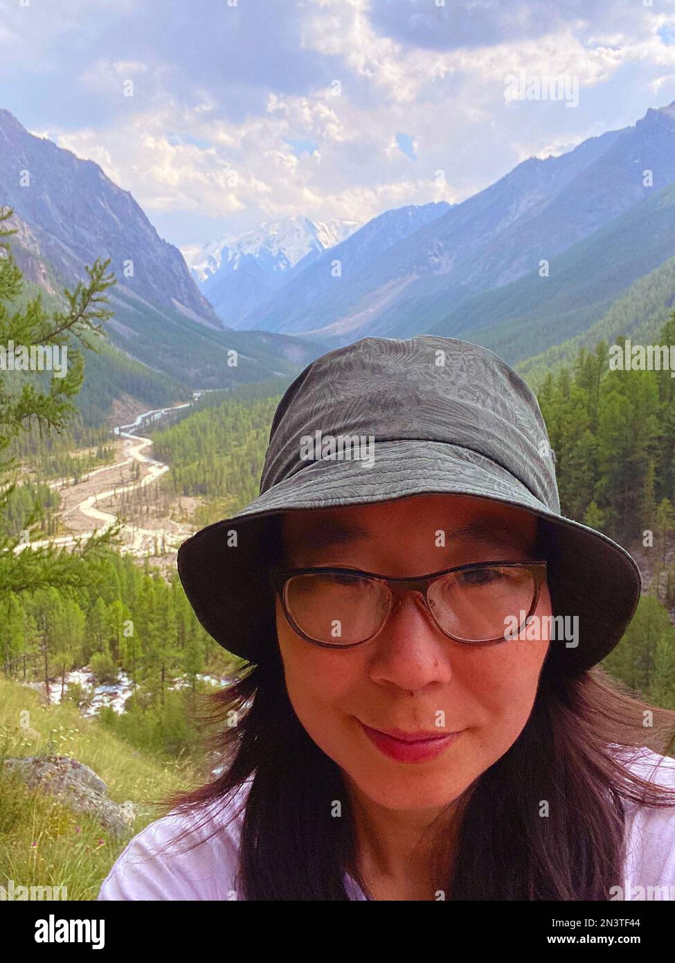 A traveler girl in glasses and a hat takes a selfie against the backdrop of a panorama of mountains and a river with glaciers and snow in Altai. Stock Photo