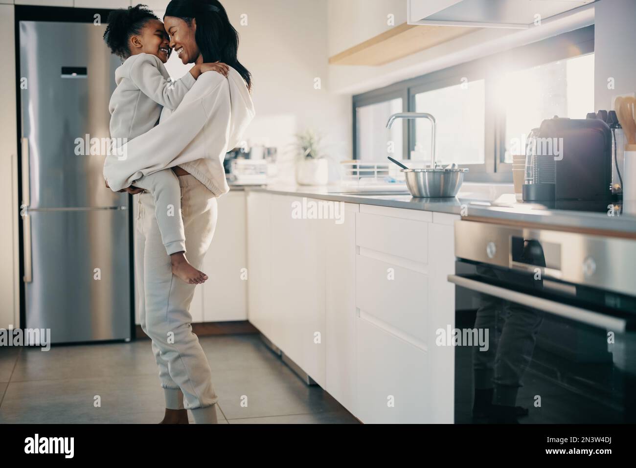 Single mother carrying her daughter in the kitchen. Happy black mom bonding with her kid. Mom and daughter are smiling and embracing each other. Stock Photo