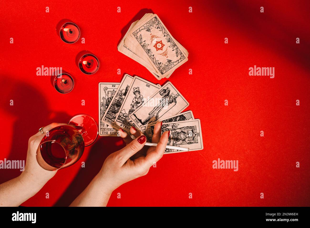 Esoteric composition top view with female hands of fortune teller with cigarette and glass of red wine reading Tarot cards on red background with Stock Photo