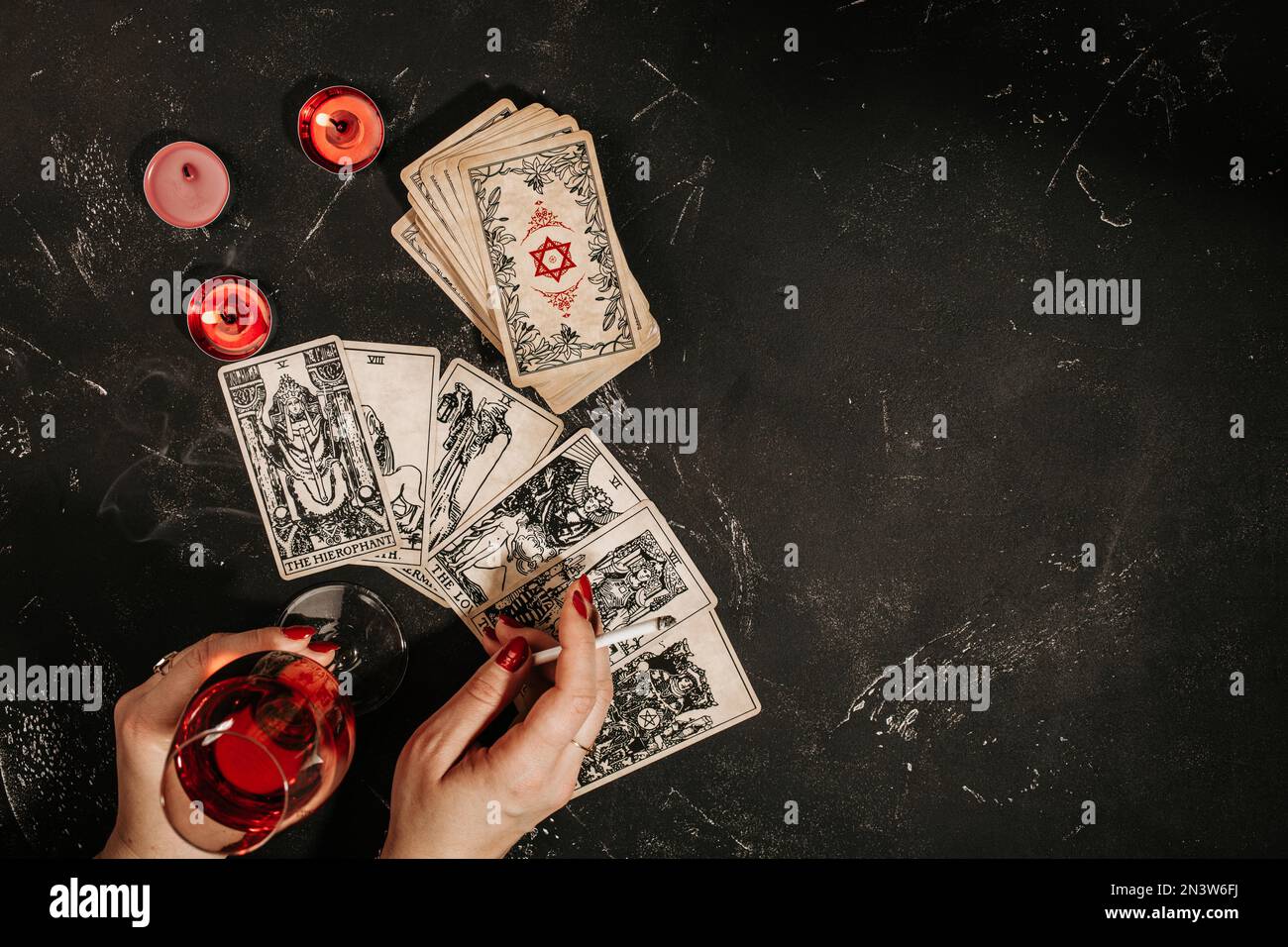 Esoteric composition top view with female hands of fortune teller with glass of red wine and cigarette reading Tarot cards on black magic table with Stock Photo
