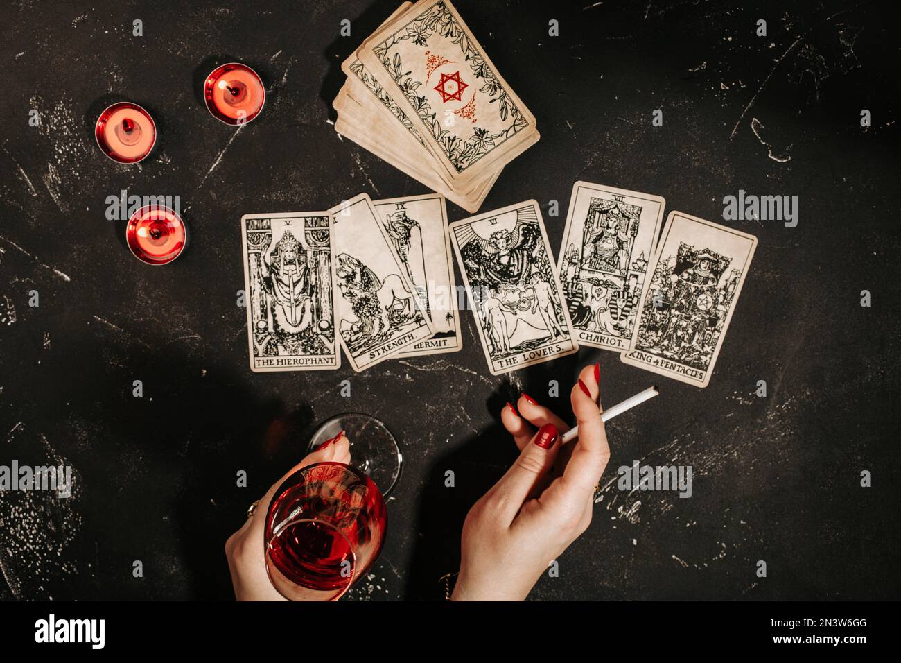 Esoteric composition top view with female hands of fortune teller with glass of red wine and cigarette reading Tarot cards on black magic table with Stock Photo
