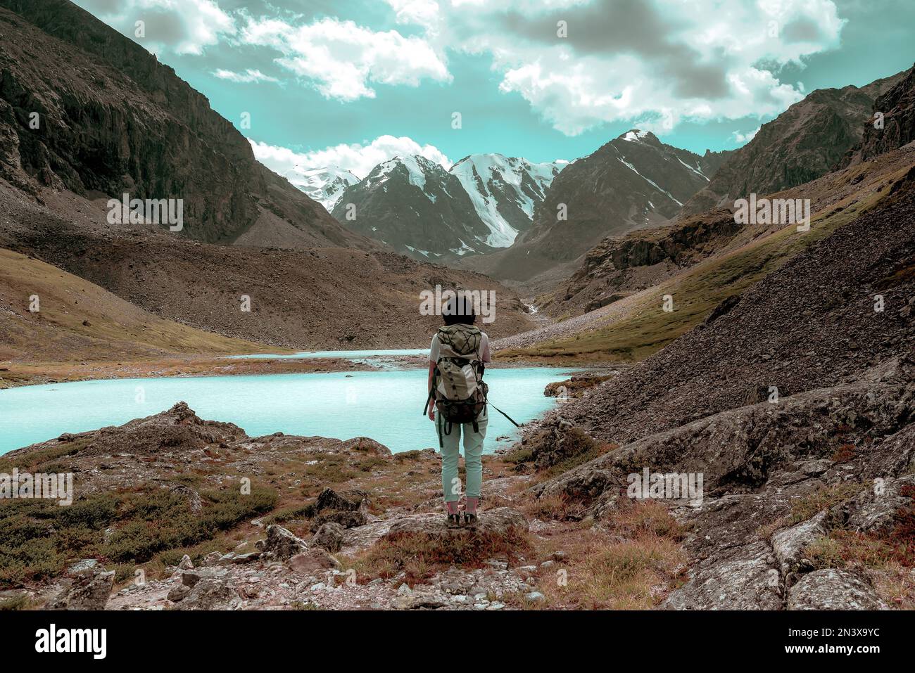 A tourist girl with a large backpack stands near an alpine lake in the stones in the Altai mountains against the backdrop of glaciers and snow in autu Stock Photo