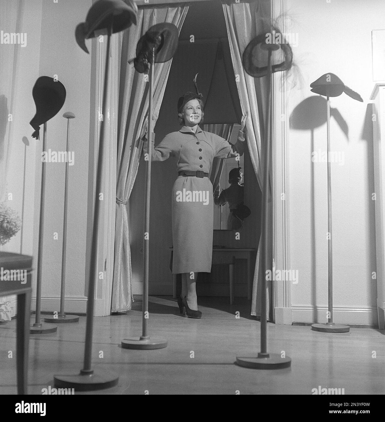 Women's fashion in the 1940s. A young woman in the opening of the fitting room wearing a typical 1940s outfit with a matching hat. In front of her are different hats mounted on stands. Sweden 1949 Kristoffersson ref AT37-4 Stock Photo