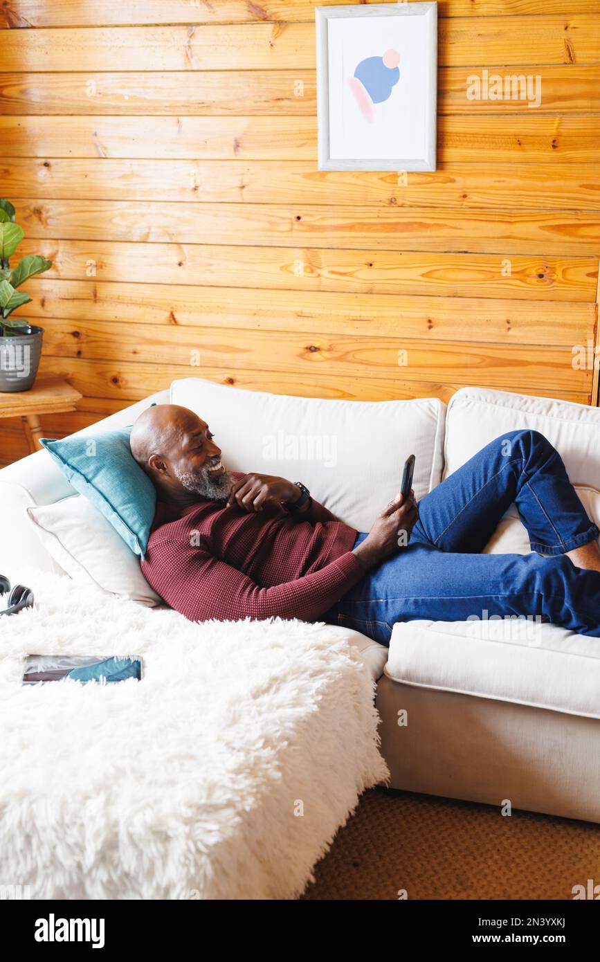 Smiling bald african american senior man using cellphone while relaxing on couch at log cabin Stock Photo
