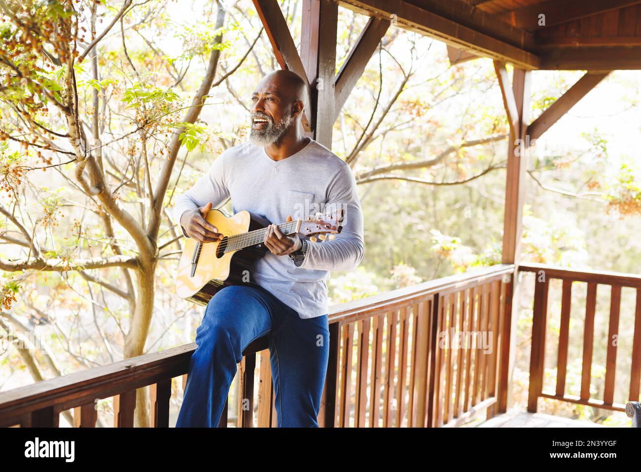 Bald african american senior man playing guitar and singing while sitting on railing in balcony Stock Photo