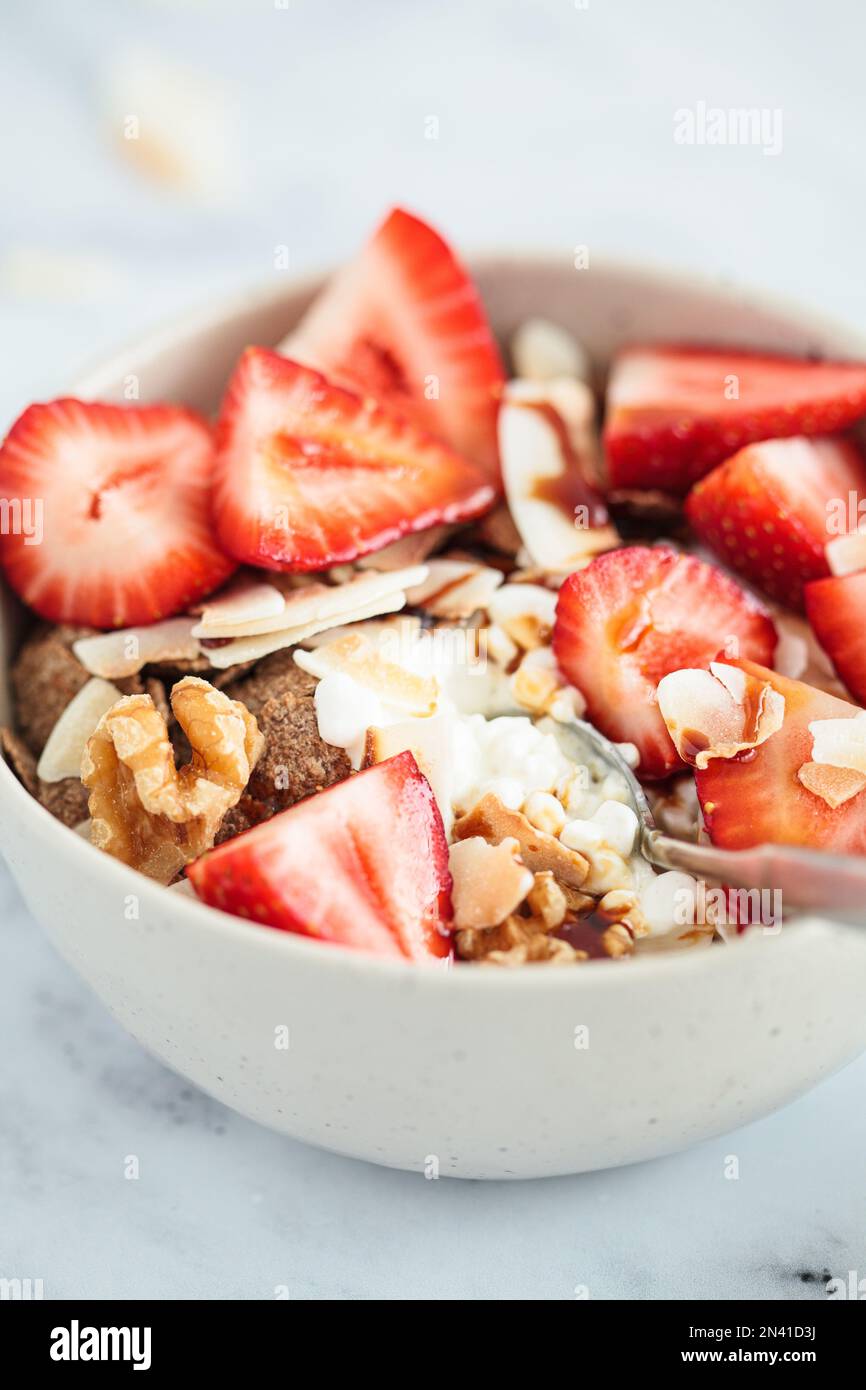 Cottage cheese bowl with strawberries, cereal flakes, coconut, nuts and syrup, white marble background, close-up. Stock Photo