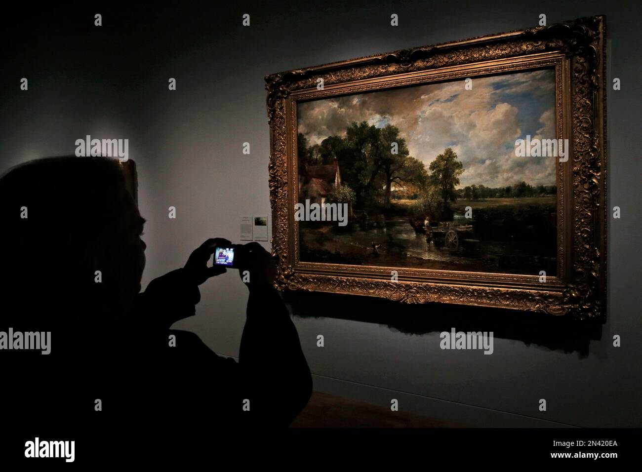 A woman takes pictures of "The Hay Wain" by British landscape painter John Constable, during a preview of an exhibition entitled: 'Constable: The Making of a Master ' with works by the artist at the Victoria and Albert Museum, in London, Wednesday, Sept. 17, 2014. Best-known to many for ‘The Hay Wain’ - an artwork which adorns countless decorative plates and trays, the exhibition explores Constable’s influences and takes a look at the creative process behind some of his most famous works. The exhibition will run from Sept. 20, 2014 to Jan. 11. 2015.(AP Photo/Lefteris Pitarakis) Stock Photo