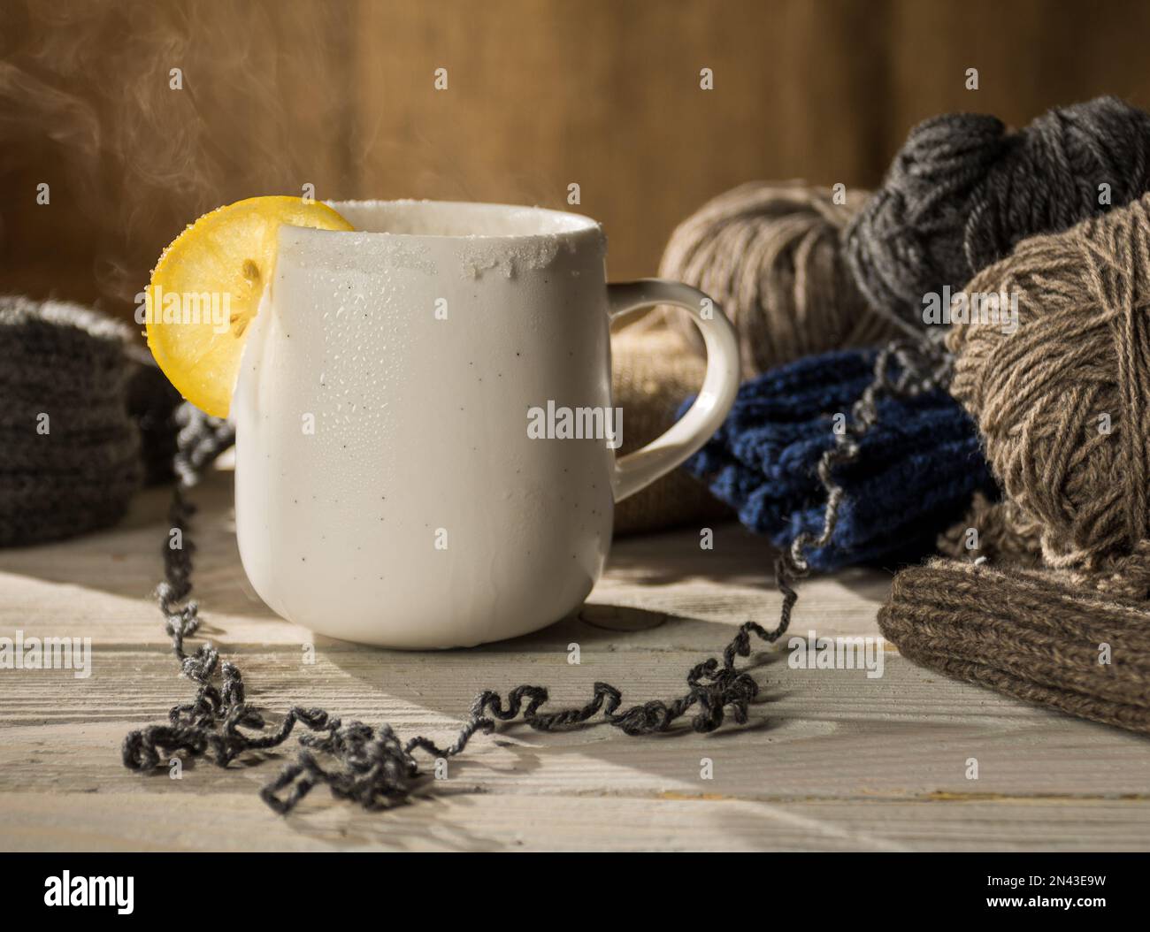 Cozy home scene - a mug of hot tea with lemon and steam on a wooden table surrounded by balls of woolen threads. Stock Photo