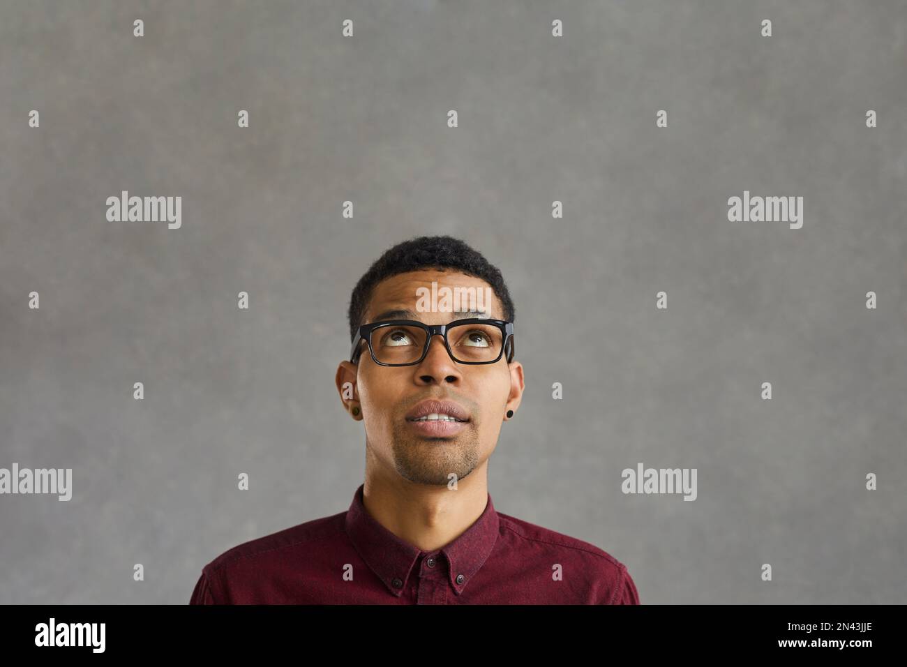 Dark-skinned man with doubtful facial expression looking up on gray concrete background. Stock Photo