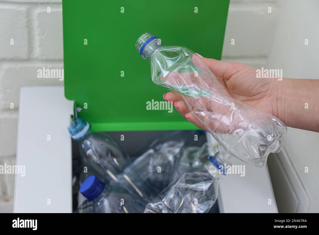 Woman putting used plastic bottle into trash bin near white brick wall ...