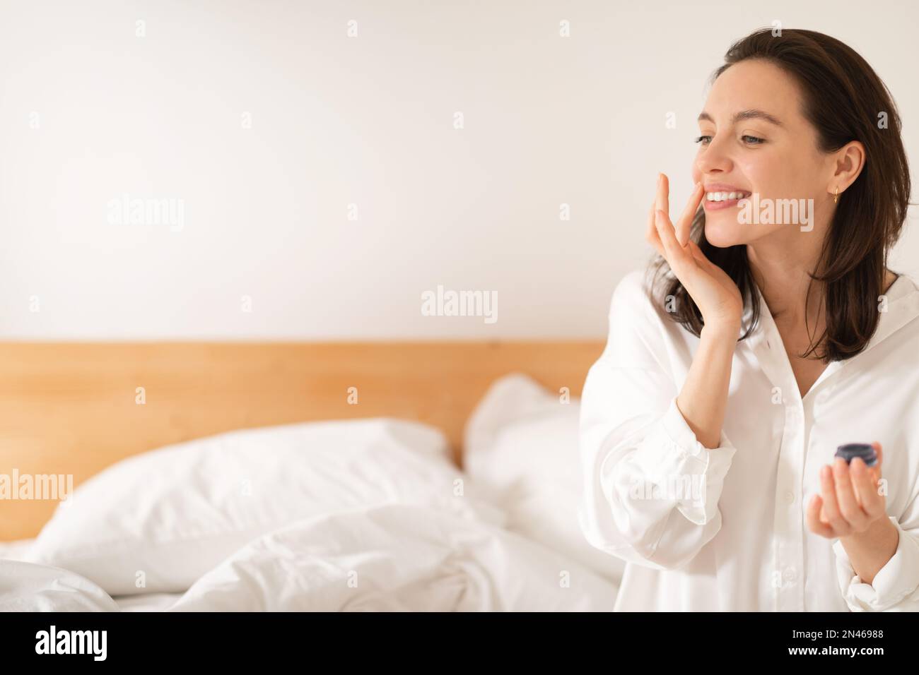 Cheerful caucasian millennial woman sitting on bed, applying moisturizing lipstick on lips, enjoy good morning Stock Photo