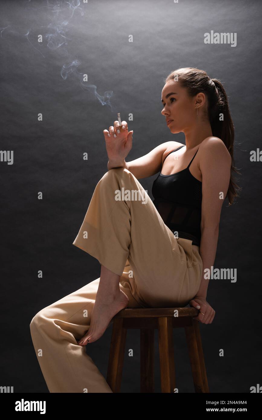Barefoot Woman In Beige Pants Holding Cigarette While Sitting On High Chair On Black Background