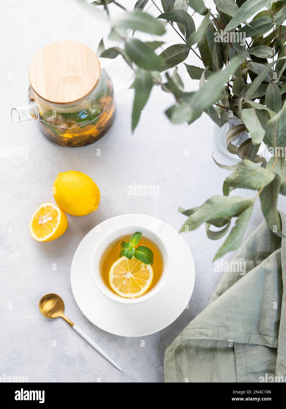 Herbal tea with lemon and mint in a white cup on a wooden plate on a light background with eucalyptus branches and a teapot. The concept of a healthy Stock Photo