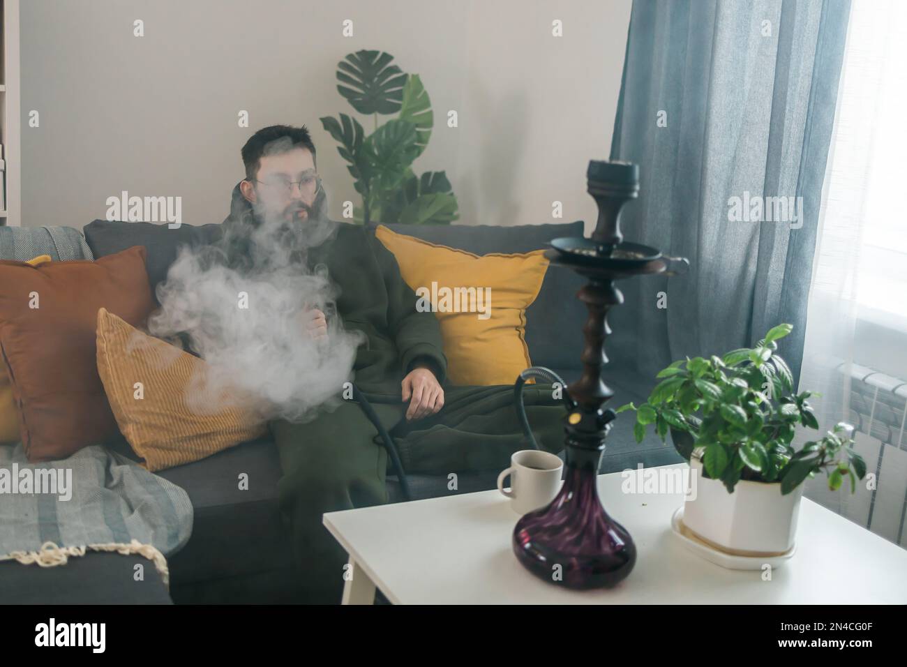 Bearded millennial or gen z man smoking hookah while relaxing on sofa