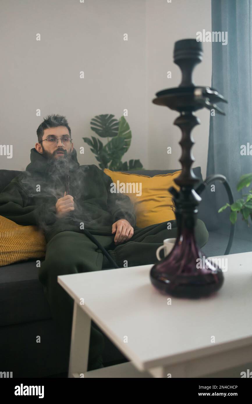 Bearded millennial or gen z man smoking hookah while relaxing on sofa