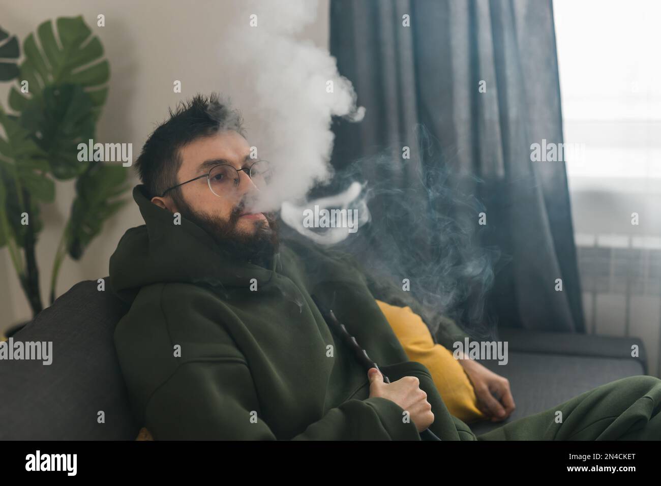 Bearded millennial or gen z man smoking hookah while relaxing on sofa