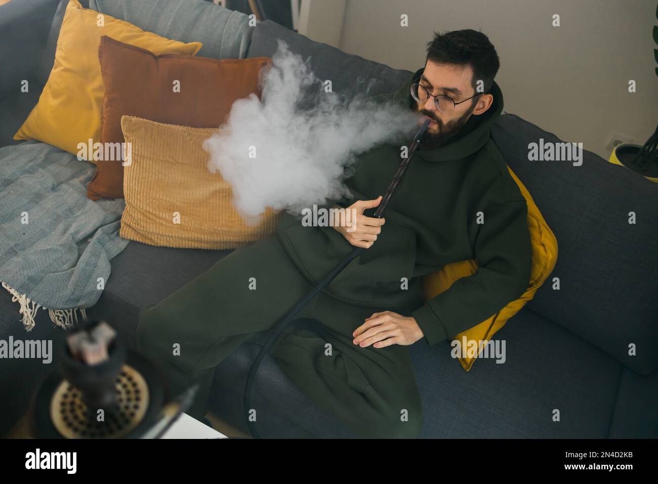 Bearded millennial or gen z man smoking hookah while relaxing on sofa