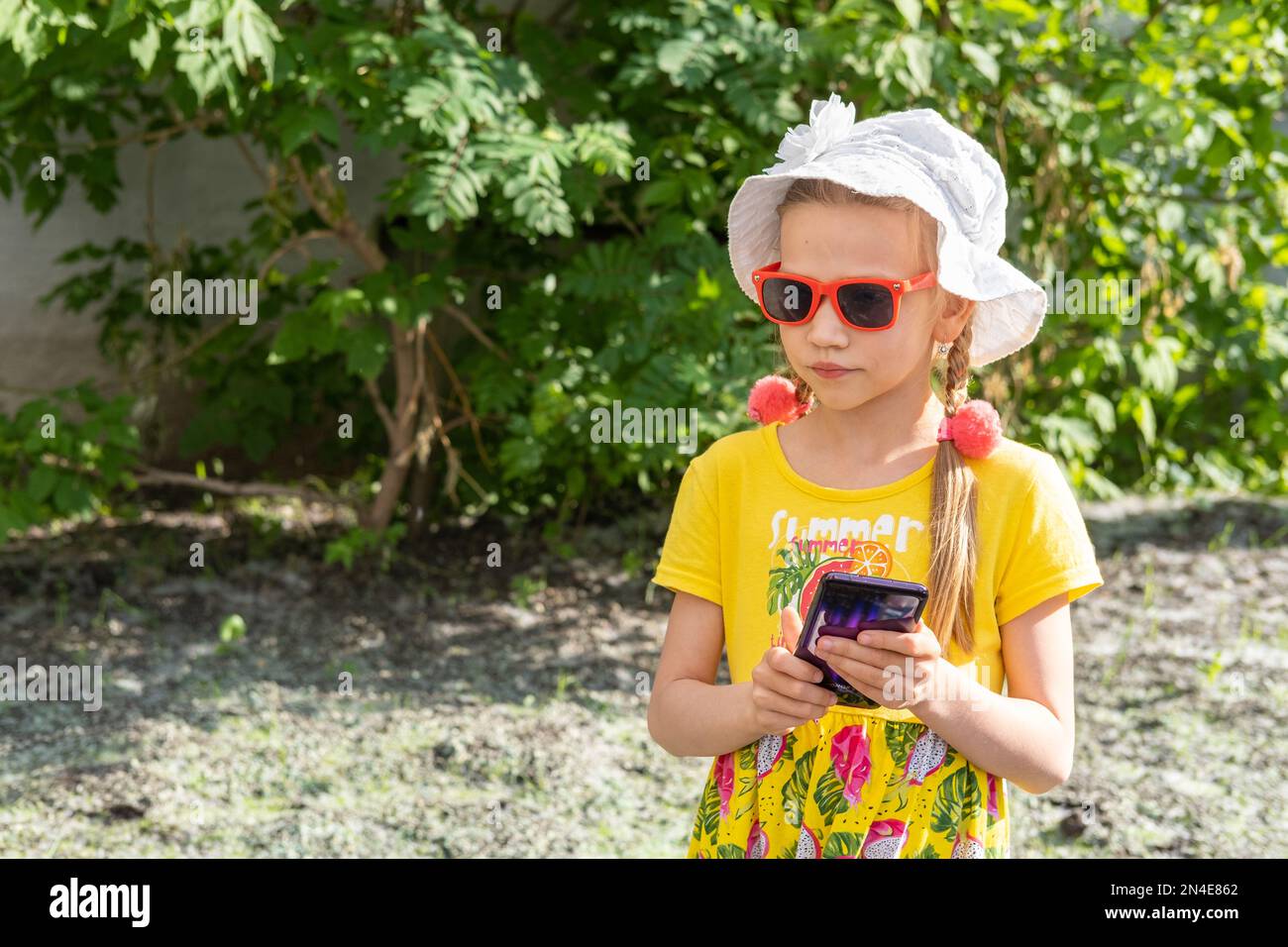 Happy smiling child girl looking in mobile phone outdoors in summer. A cheerful little girl in a yellow dress, a white hat and sunglasses holds a smar Stock Photo