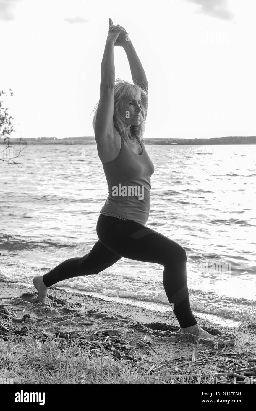 An elderly woman is doing sports on the lawn on the shore of the lake. The woman stands in the warrior pose with her hands up. Yoga pose. Outdoor spor Stock Photo