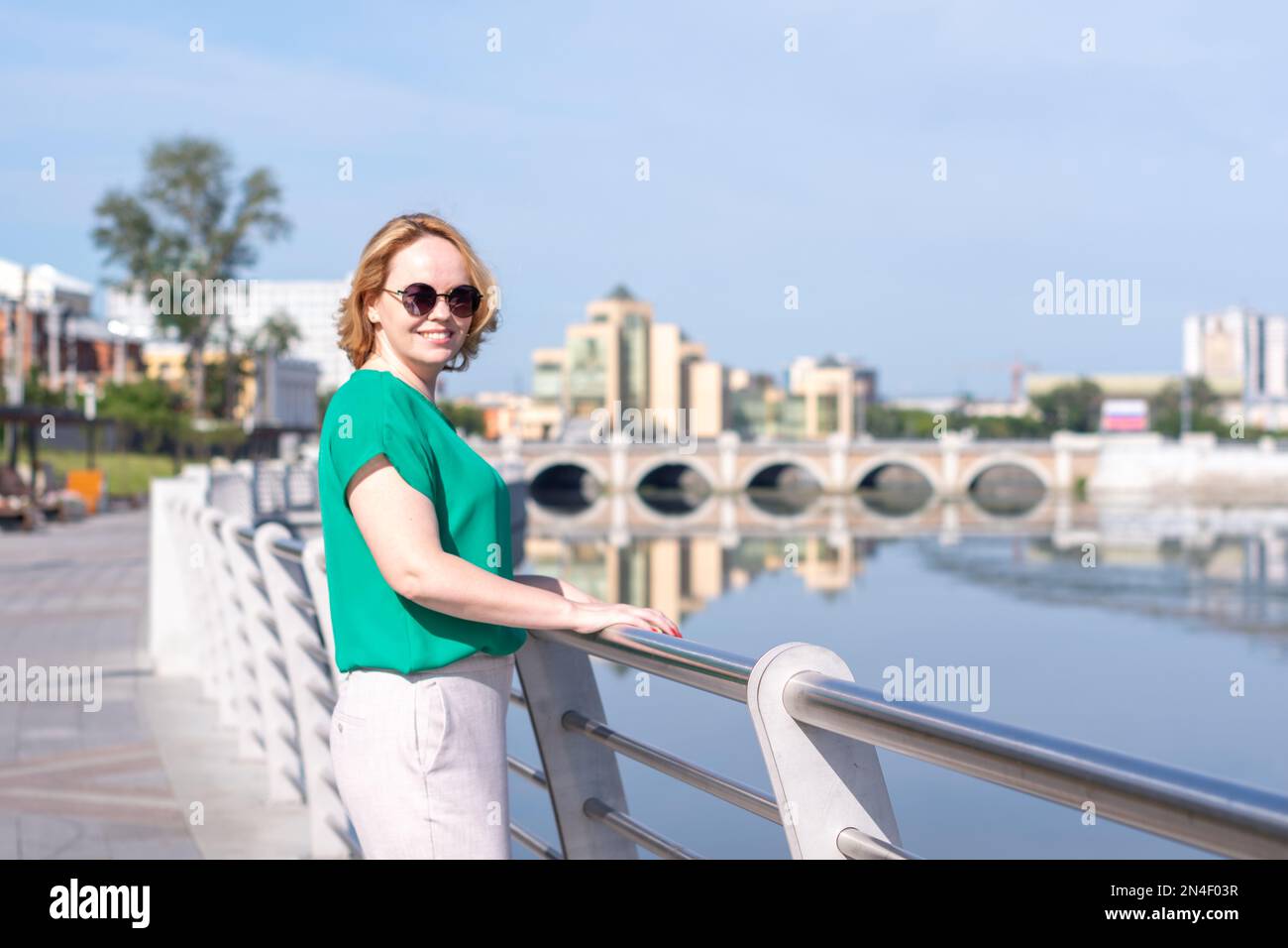 Portrait of a girl standing on the bank of the river holding on to the fence against the background of a blurred city. A girl walks through streets of Stock Photo