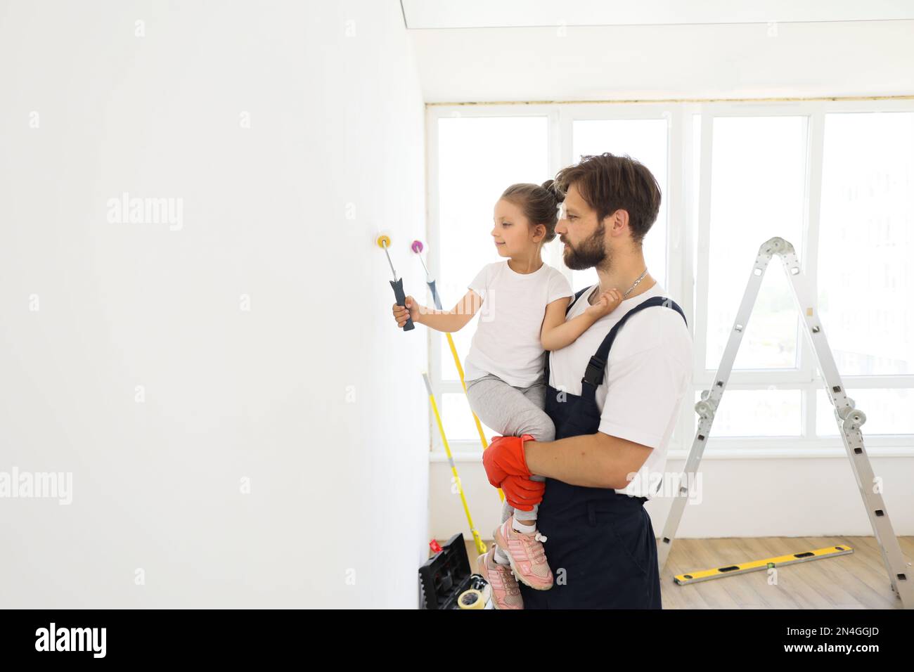 Smiling 5-year-old girl holds a paint roller in her hands. The child helps her father, who holding her, near the wall, in the repair. Stock Photo
