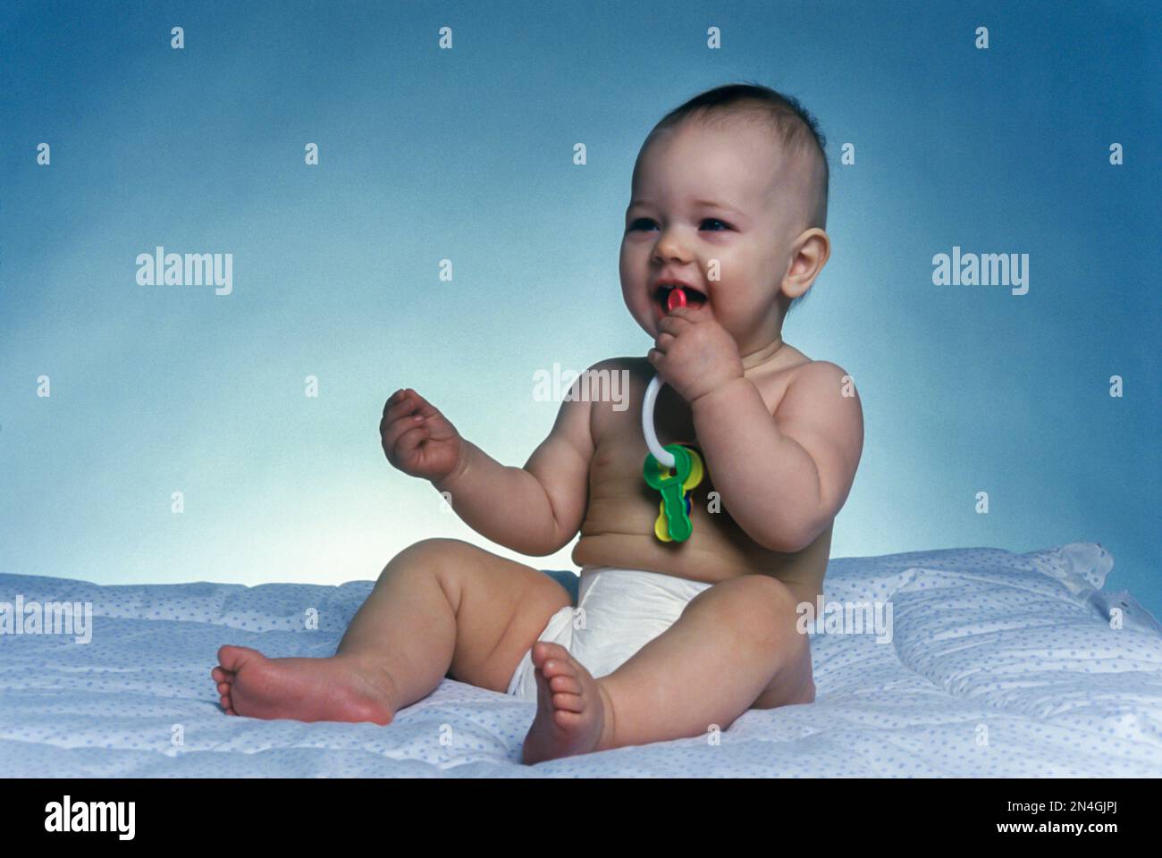 CAUCASIAN INFANT BABY SITTING PLAYING WITH PLASTIC TOY KEYS ON WHITE BLANKET Stock Photo