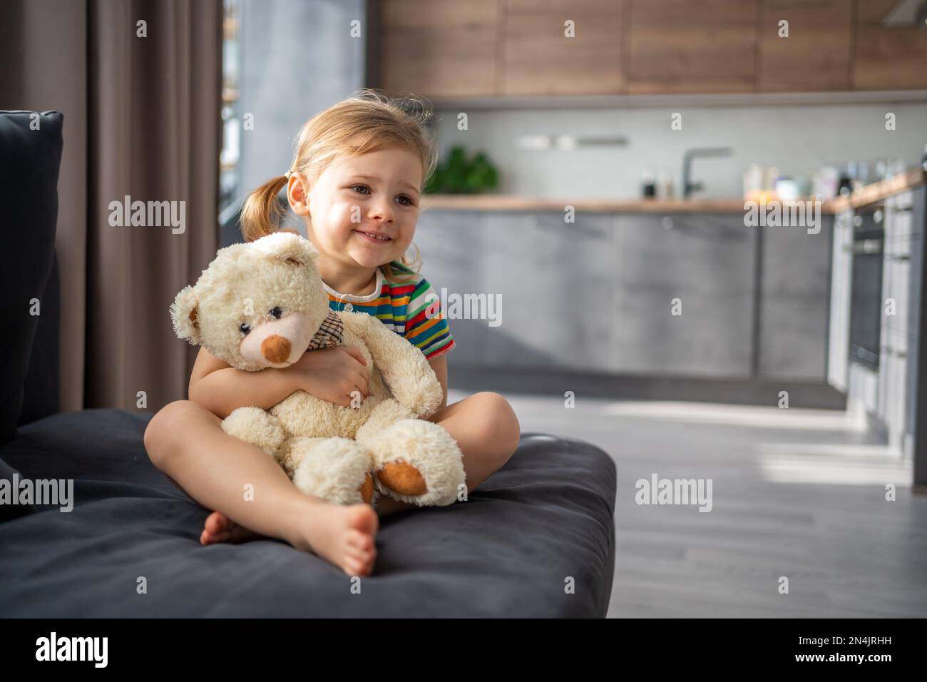 Cute little girl hugging teddy bear while sitting on sofa at home Stock ...