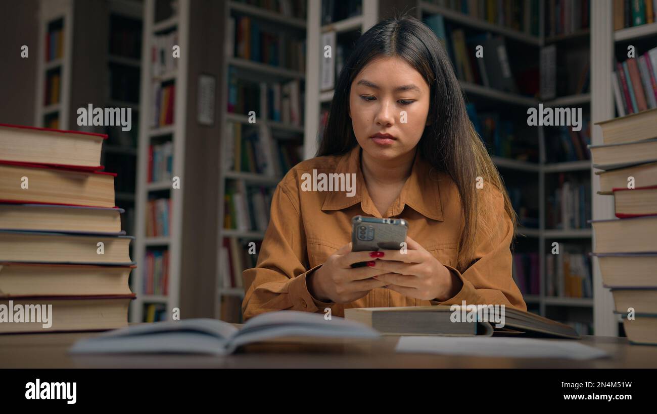 Young serious asian woman student sitting in library at desk searching information on Internet using mobile phone preparing for exam use free wifi on Stock Photo