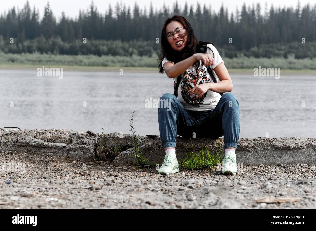 Asian girl-traveler on the edge of an abandoned stone pier throws a stone forward grimacing and having fun on the background of the Northern forest. Stock Photo