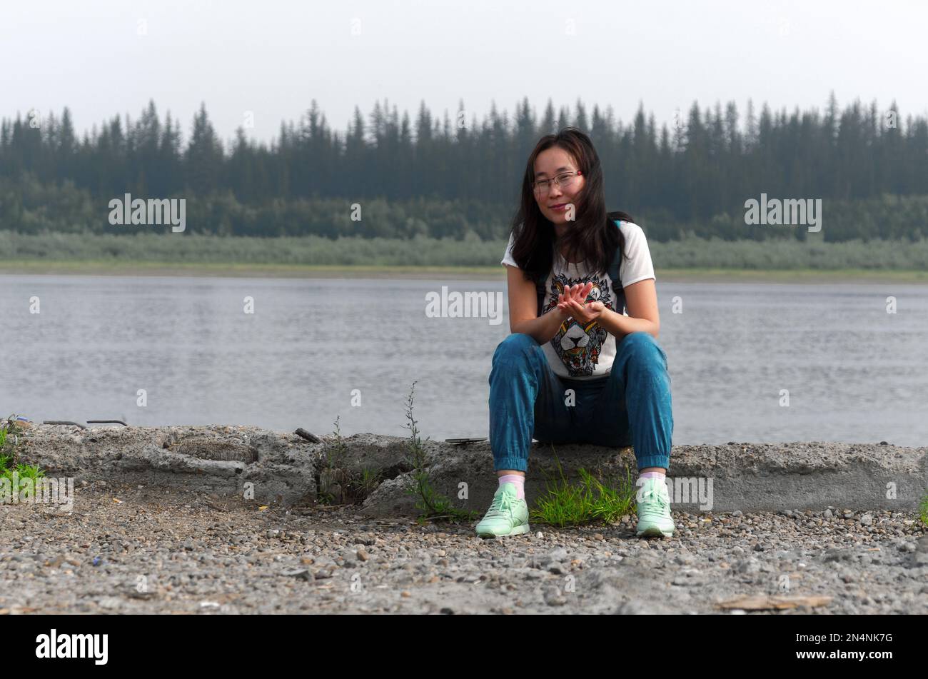Smiling girl-traveler Asian girl is sitting on the edge of the old pier on the river Vilyui forests of Yakutia in the far North of Russia slapping in Stock Photo