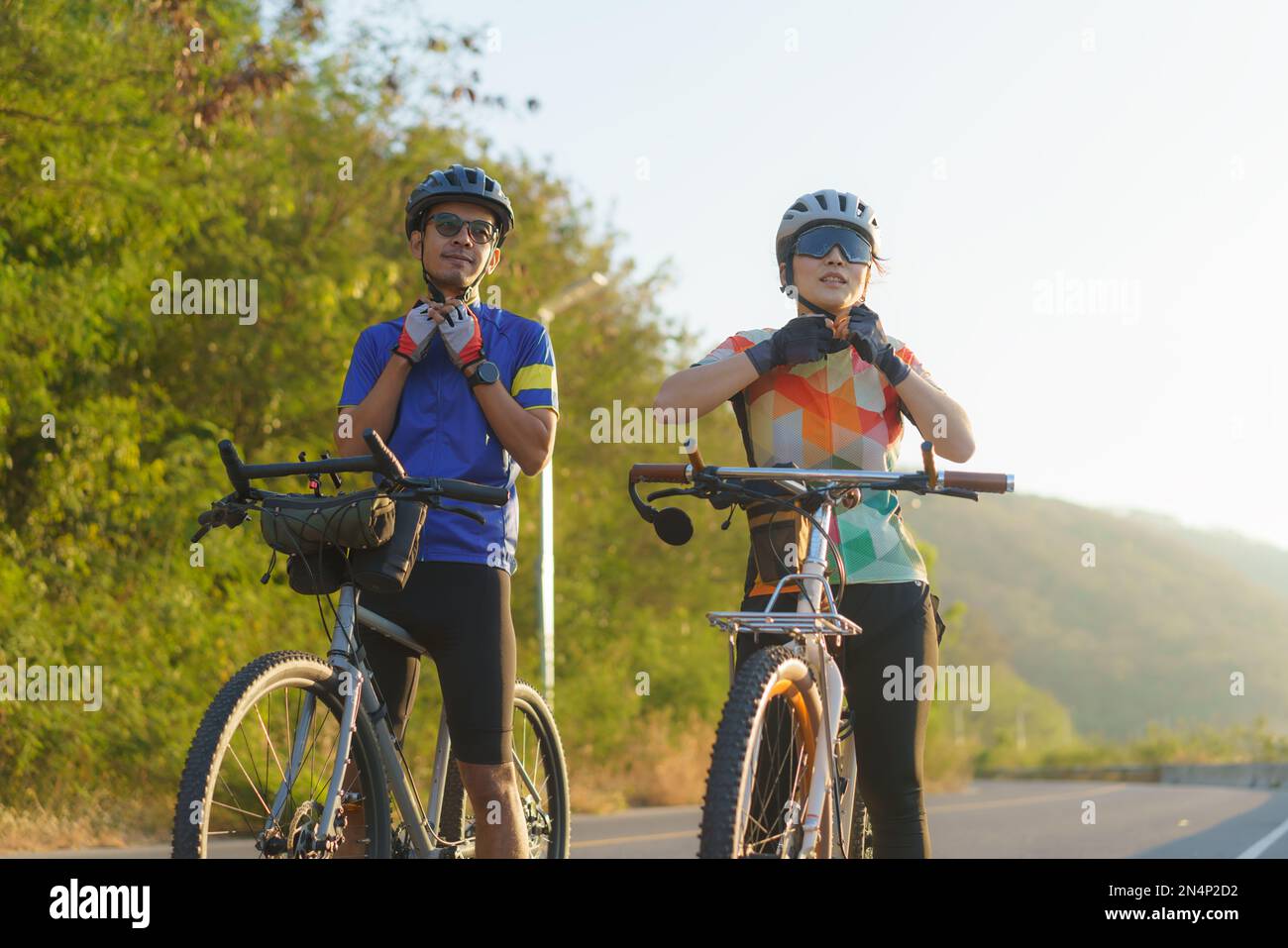 Asian cyclist couple dons helmets and chin straps as they prepare for their bike ride around a beautiful morning lake. Stock Photo
