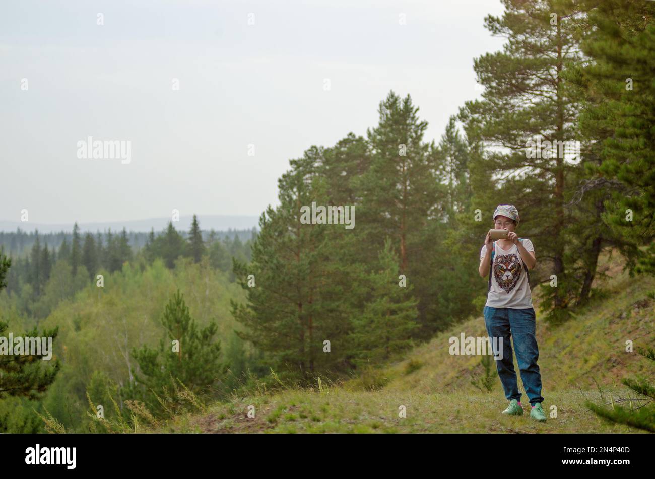 Yakut Asian girl tourist takes pictures on the phone view from the mountain in the spruce forest of Northern Yakutia with forest to the horizon. Stock Photo
