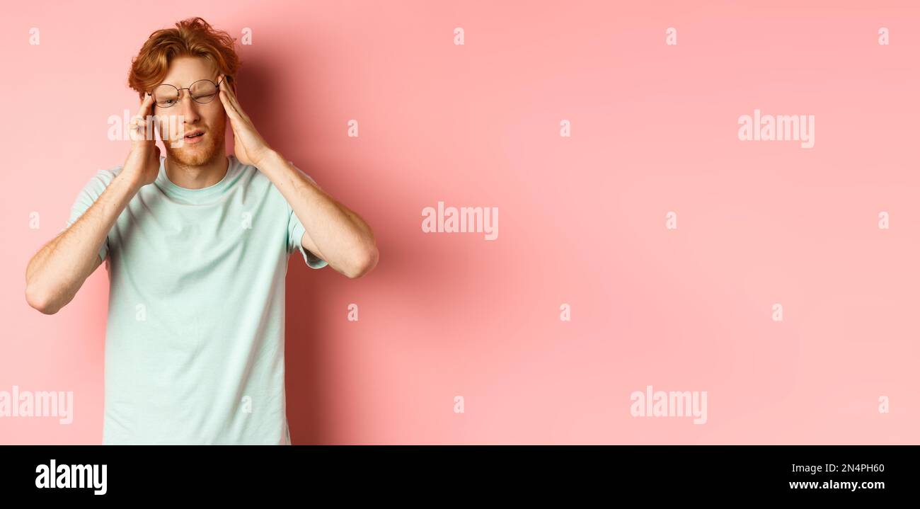 Portrait of redhead man in crooked glasses touching head and feeling dizzy or nauseous, having hangover or headache, standing over pink background Stock Photo